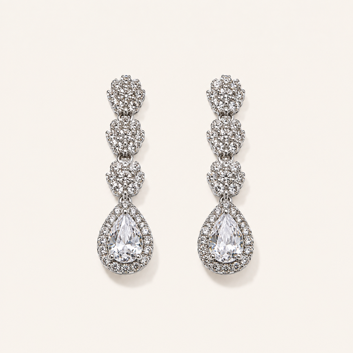 A pair of silver-tone drop earrings adorned with clear pavé gemstones, featuring a stacked floral design and a teardrop-shaped center stone, displayed on a light cream background.