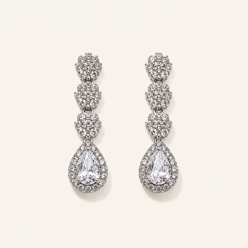 A pair of silver-tone drop earrings adorned with clear pavé gemstones, featuring a stacked floral design and a teardrop-shaped center stone, displayed on a light cream background.