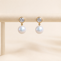 A pair of gold drop earrings with clear round gemstones at the top and large white pearl-like drops below, displayed on a beige jewelry stand against a soft cream background.