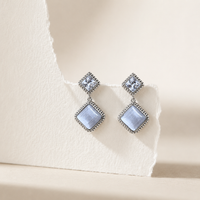 A pair of silver-toned drop earrings with square top studs and dangling diamond-shaped charms, photographed on a warm cream textured surface with torn-paper styling.