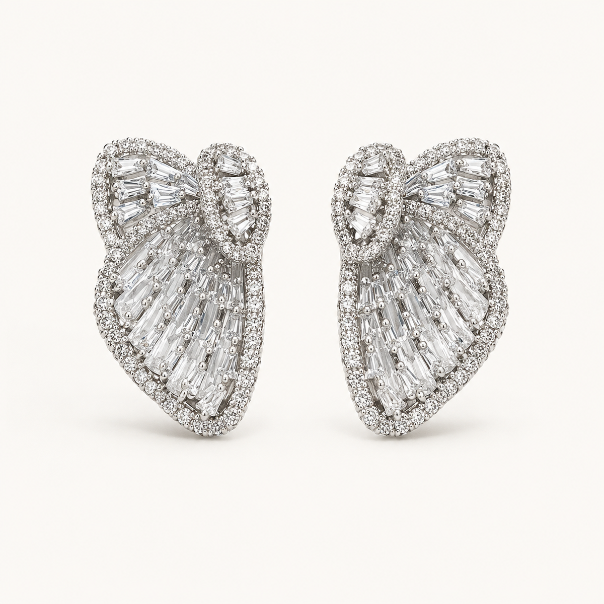 A pair of large bow-shaped stud earrings set with clear baguette and round-cut gemstones in a silver-toned metal, photographed on a clean off-white background.
