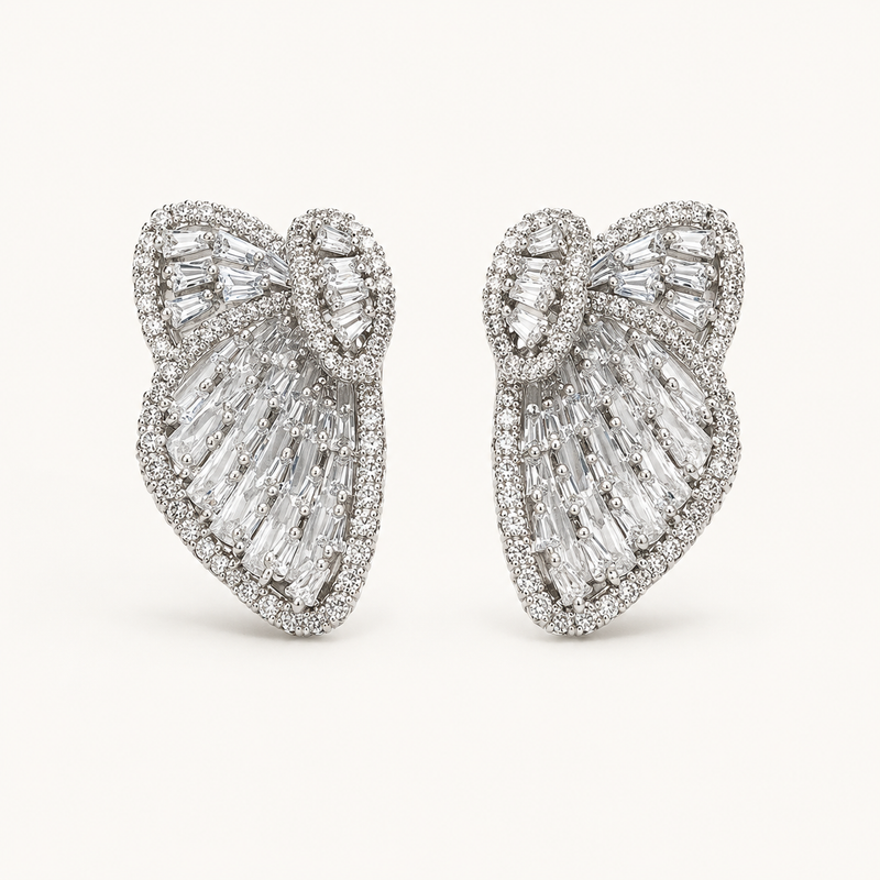 A pair of large bow-shaped stud earrings set with clear baguette and round-cut gemstones in a silver-toned metal, photographed on a clean off-white background.