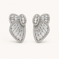 A pair of large bow-shaped stud earrings set with clear baguette and round-cut gemstones in a silver-toned metal, photographed on a clean off-white background.
